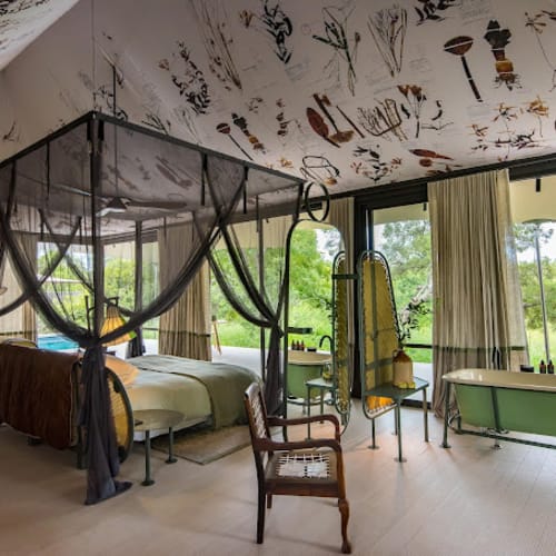 Interior of Saseka Tented Camp with a four-poster bed, armchair, and large windows showing green landscape.