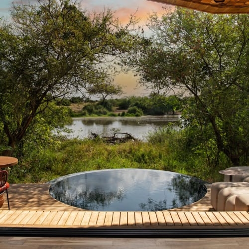 A luxurious lodge room with a round plunge pool overlooking a river, surrounded by lush greenery and a colorful sunset sky.