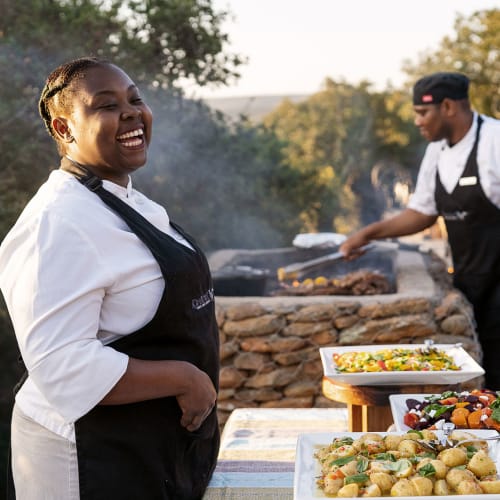 Friendly staff preparing and serving food outdoors at a scenic reserve, with a woman smiling in the foreground.