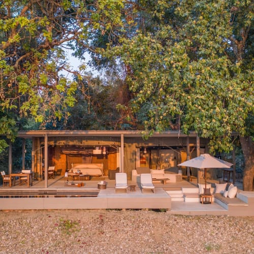 Ein modernes Safarilodge mit Sitzbereich im Freien und Liegen, auf einer leicht erhöhten Holzplattform inmitten üppiger grüner Bäume.