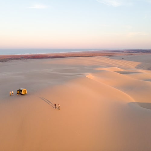 Weites Wüstenlandschaft mit rollenden Sanddünen, einem kleinen Fahrzeug und einer Person, die in der Dämmerung über die Dünen läuft.