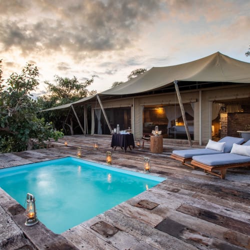An outdoor luxury suite with a small swimming pool on a wooden deck, surrounded by nature, under a partly cloudy evening sky.