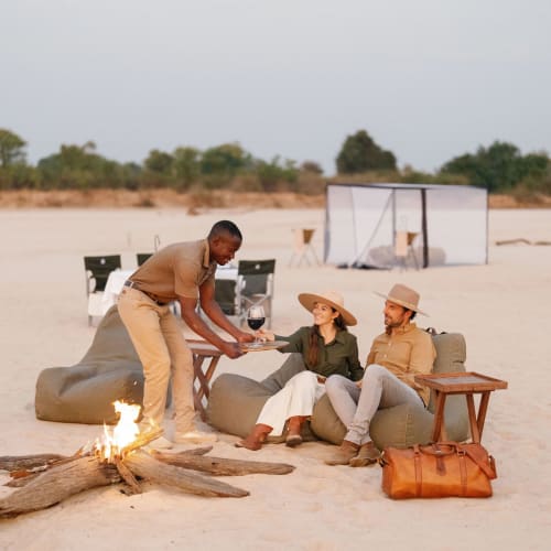 Eine Gruppe von drei Personen, die am Lagerfeuer auf dem Sandwüstenboden unter einem bewölkten Himmel sitzen, mit Liegestühlen, einem Tisch und einem Zelt im Hintergrund.