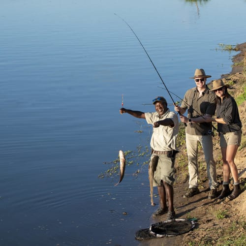 Three people stand on a riverbank, fishing and showing off a caught fish.