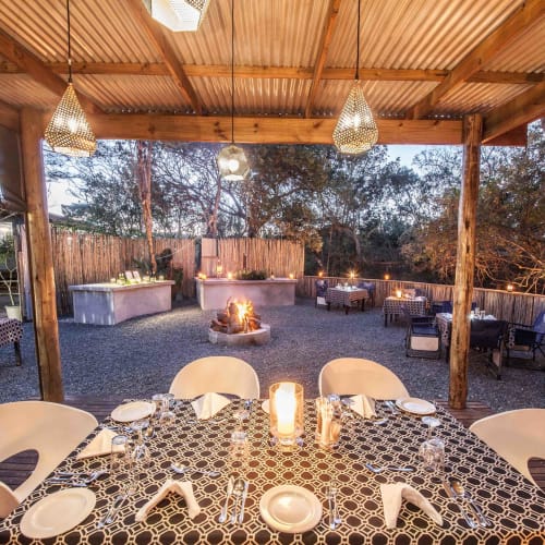 An outdoor dining area under a wooden roof with lit candles and a view of a decorated yard in the evening.