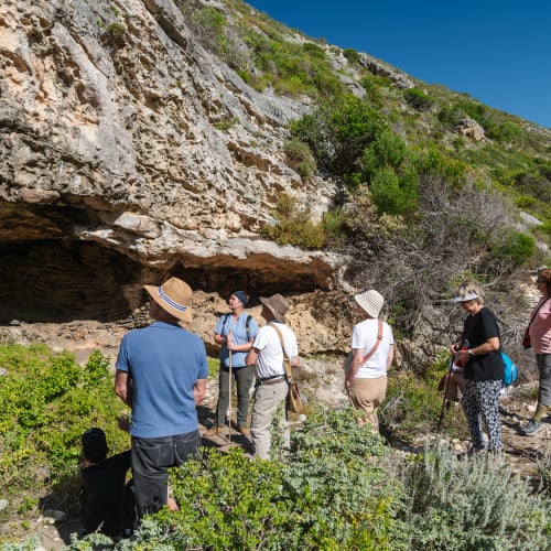 Gruppe von Menschen auf einer Wanderung in einer natürlichen Umgebung mit Felsen und Vegetation, einige mit Hüten und Wanderstöcken, die eine Höhle in einer grünen Klippe unter einem klaren Himmel beobachten.
