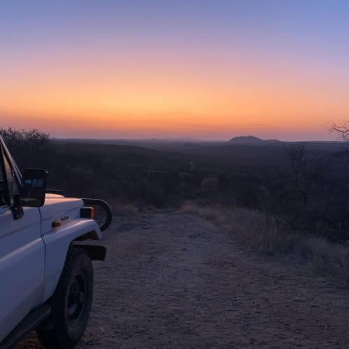 A white vehicle parked on a dirt path observing a colorful sunset with a landscape of sparse trees and distant hills.