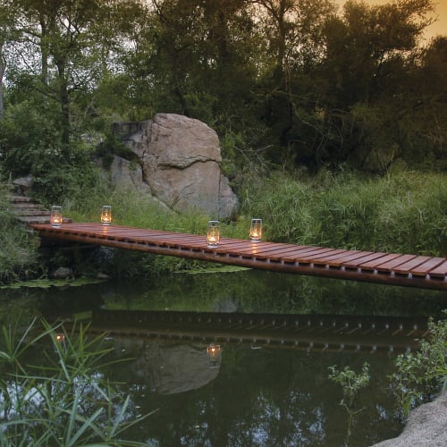 Eine kleine Holzbrücke über einem ruhigen Wasserpool, umgeben von Grünflächen und Felsen, mit Laternen entlang der Brücke bei Sonnenuntergang.