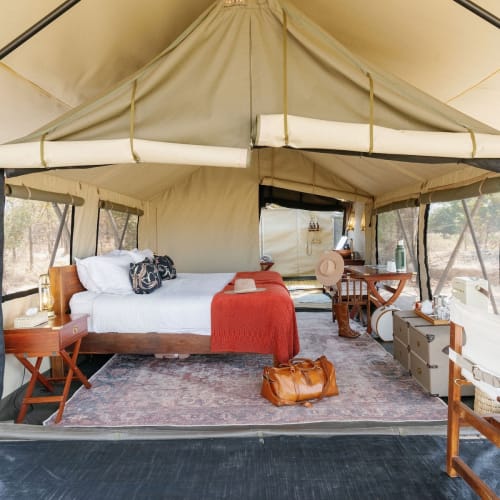 Interior of a luxurious tent set up as a cozy lodge with a bed, armchair, and wooden furniture, surrounded by nature.