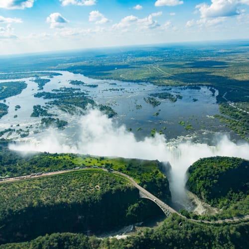 Luftaufnahme von den Victoria Falls mit dem Wasserfall, der die Landschaft überspannt, und einer Brücke, die den Zambezi-Fluss überquert.