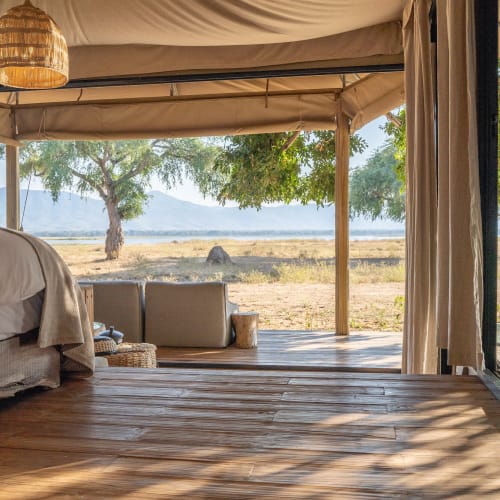 Ein Safari-Zelt mit Blick über eine afrikanische Landschaft, mit einem Bett und Sitzecke im Freien unter einem Vordach.