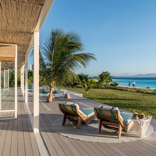Luxury villa veranda overlooking a beach with palm trees and clear blue sea under a bright sky.