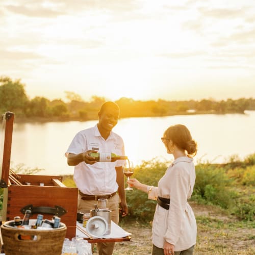 Ein Mann und eine Frau genießen gemeinsam ein Getränk im Freien am Fluss bei Sonnenuntergang, mit einer kleinen tragbaren Bar und einer natürlichen Kulisse.