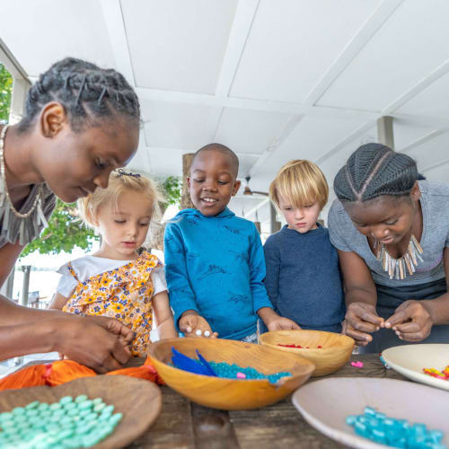 Eine Gruppe vielfältiger Kinder und zwei Frauen beschäftigen sich mit einer kreativen Aktivität mit bunten Perlen und Dekorationen auf einem Holztisch.