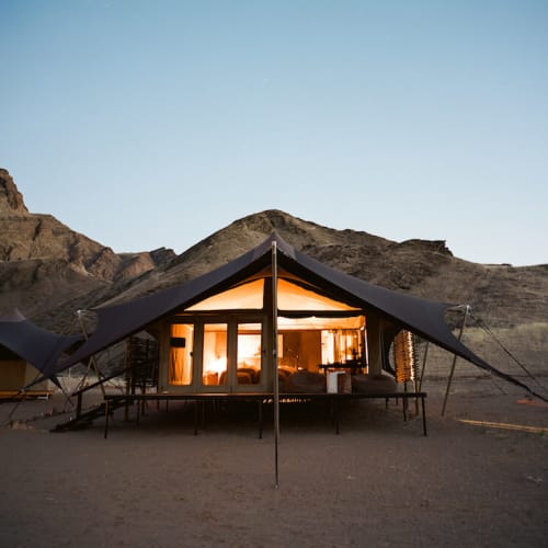 A large camping tent set up in a desert landscape with mountains in the background, illuminated from within at dusk.