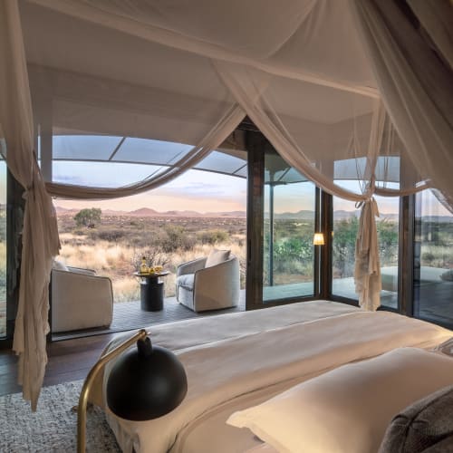 A bedroom with a view of the Kalahari landscape through large glass windows, featuring a bed and outdoor seating area.