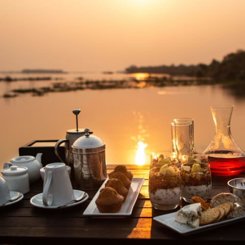 A breakfast setup on a wooden table near a river at sunset, with cups, a teapot, desserts, and a drink surrounded by a scenic landscape.