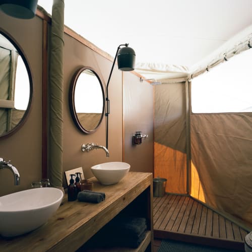 A rustic bathroom with two circular mirrors, wooden countertop with two vessel sinks, and a tent window in Namibia.