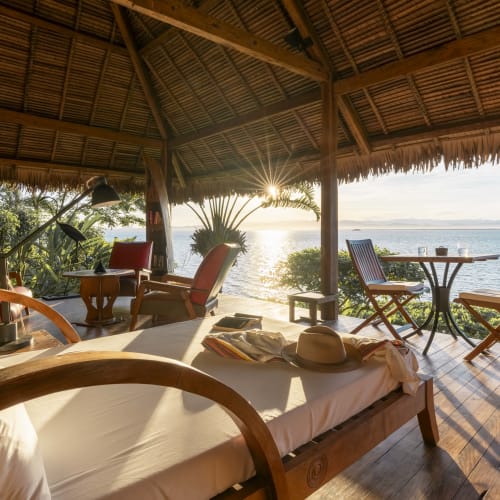 Eine luxuriöse Holzterrasse mit Blick auf den Ozean, ausgestattet mit einem Bett, Stühlen und einem Tisch, umgeben von dichtem Grün.