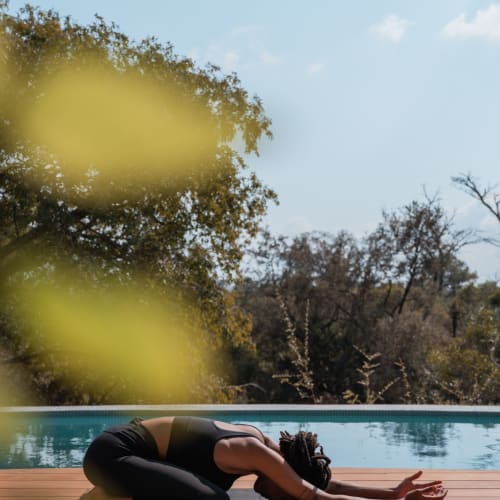 A person practicing yoga outdoors near a pool with trees in the background on a sunny day.