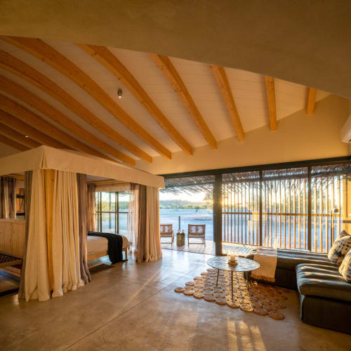 Innenansicht einer geräumigen Suite mit einem vierpostigen Bett und einem Sitzbereich, beleuchtet durch warmes Sonnenlicht durch Glas türen mit Blick auf ein Gewässer.
