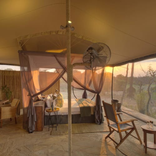 Ein luxuriöses Safarizelt mit Bett, Sitzecke und großen Fenstern, die eine Naturlandschaft im Sonnenuntergang zeigen.