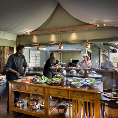 Gäste und Personal in einer offenen Küche bei Duba Plains, Botswana, mit Köchen beim Essen und Besuchern im Gespräch.