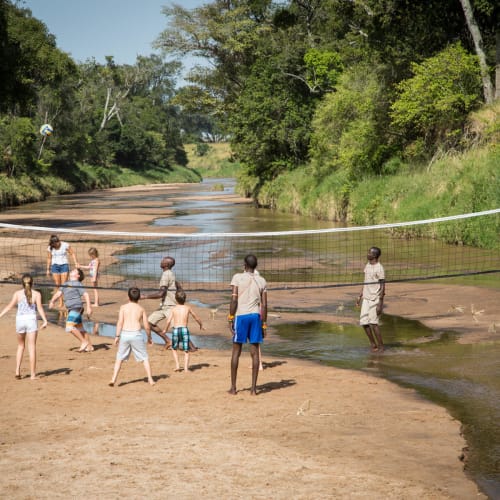 Children and adults playing volleyball on the sandy riverbank in Salas Camp, surrounded by lush green trees.