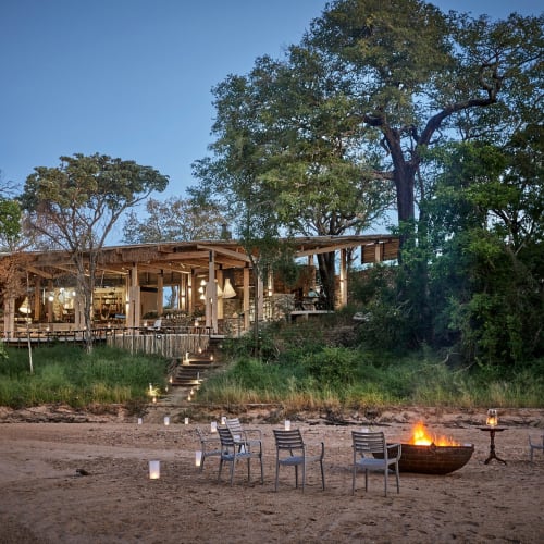 A rustic tented camp set in a natural environment with a beach and a bonfire surrounded by chairs.