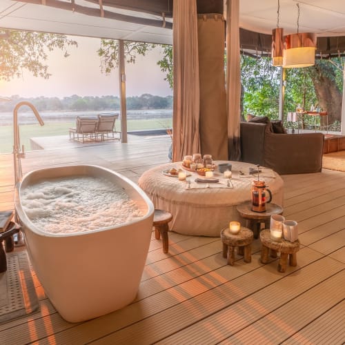 Luxuriöses Zelt im Open-Air-Design mit Badewanne, kleinen Holzscheiten mit Kerzen und Blick auf den Fluss beim Sonnenuntergang.