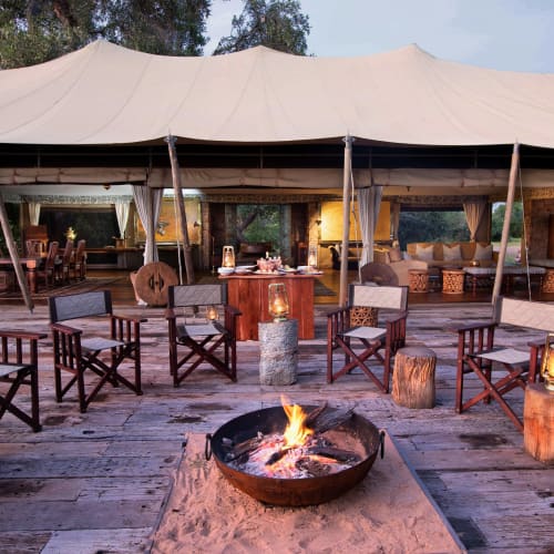 A cozy outdoor seating area with wooden chairs arranged around a campfire under a large tent, with lanterns on stumps, in a wilderness setting.