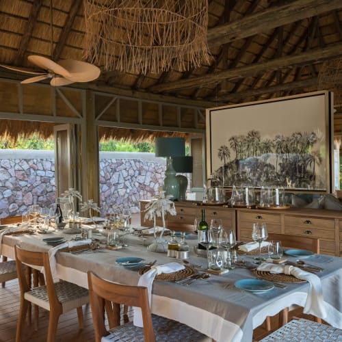 A luxurious indoor dining area with a long table set for a meal, surrounded by wooden chairs, with large vases, artwork, and natural materials decorating the space.