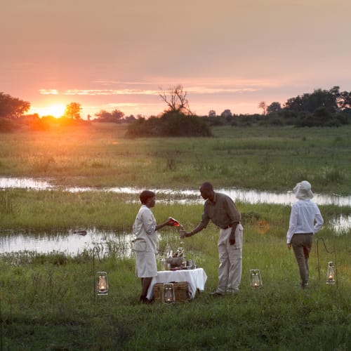 Three people enjoying a sundowner picnic on a grassy plain at sunset in Botswana, with lanterns and a small table.