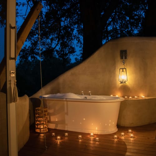 A luxurious outdoor bathtub setup on a wooden deck at night, illuminated by candles and a lantern, with trees and dark sky in the background.
