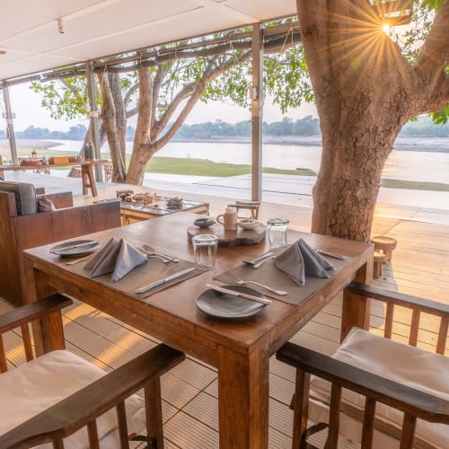 A lakeside dining setup with a wooden table, chairs, and a view of the water with trees and sunlight.