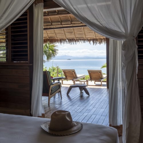 Ein Schlafzimmer mit weißen Vorhängen, die geöffnet sind und einen Balkon mit Blick auf den Ozean, Liegestühle und tropische Bäume zeigen.