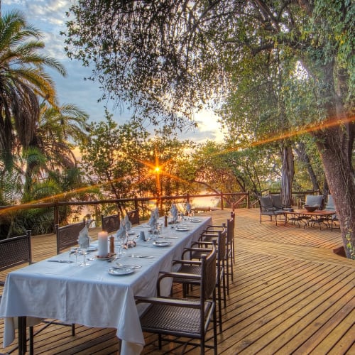 A dining table set on a wooden terrace surrounded by trees, with a sunset in the background and sunlight streaming through the leaves.