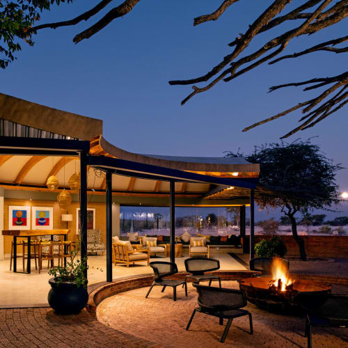 Ein Außenbereich mit Sitzgelegenheiten und Feuerstelle vor einer modernen Lodge-Struktur bei Dämmerung, mit Bäumen und Landschaft in Namibia.