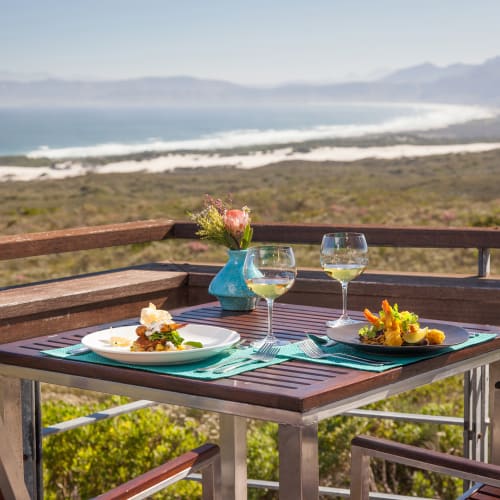 Esstisch im Freien auf einer Holzterrasse mit Blick auf eine Küstenlandschaft mit Bergen und Meer in der Ferne, gedeckt mit zwei Tellern mit Gourmetessen, zwei Gläsern Weißwein, einer Blumenvase und einem Weinkühler.