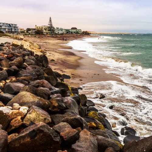 Eine felsige Küste mit den Wellen des Ozeans, die gegen das Ufer schlagen, und Gebäude im Hintergrund in Swakopmund, Namibia.