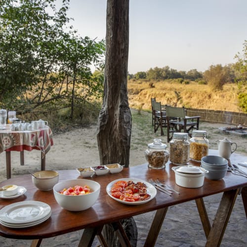 A breakfast buffet set up outdoors with various bowls, jars, and plates on a wooden table, overlooking a natural landscape with trees and chairs.