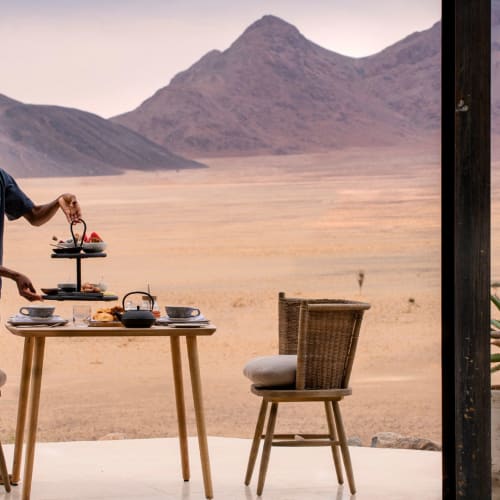 A person serving breakfast at an outdoor table with a desert landscape and mountains in the background.