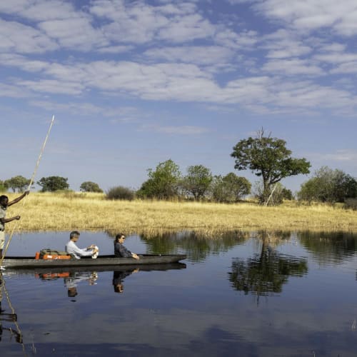 Three people on a mokoro boat navigating through a calm river in the Okavango Delta, Botswana with lush trees and a partly cloudy sky.