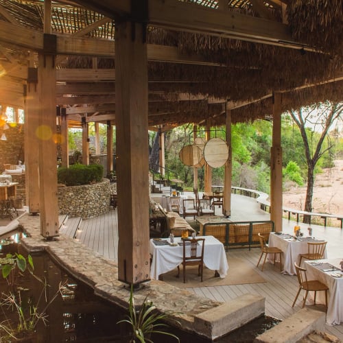 A cozy outdoor restaurant with white tablecloths and wooden chairs under a thatched roof, with trees and a sandy landscape in the background.