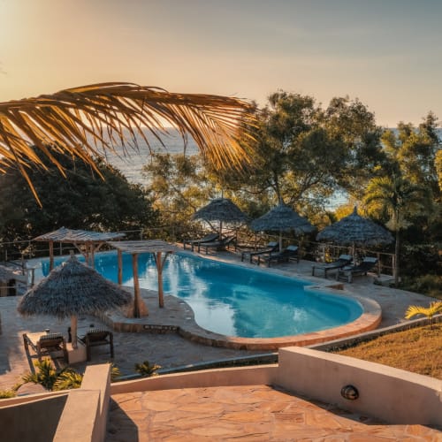 Sunset over a tropical pool area with thatched umbrellas and lounge chairs at Manta Resort, Pemba Island.