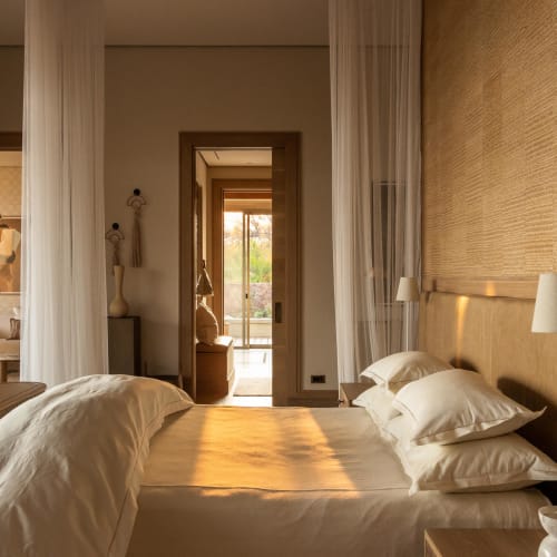 Sonnenlicht erhellt ein gemütliches Schlafzimmer mit einem ordentlich gemachten Bett, Nachttischlampen und transparenten Vorhängen, mit Blick auf einen Balkon draußen.
