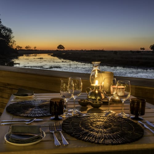 A beautifully set outdoor dinner table by a river during sunset with a lantern and candles illuminating the setting.