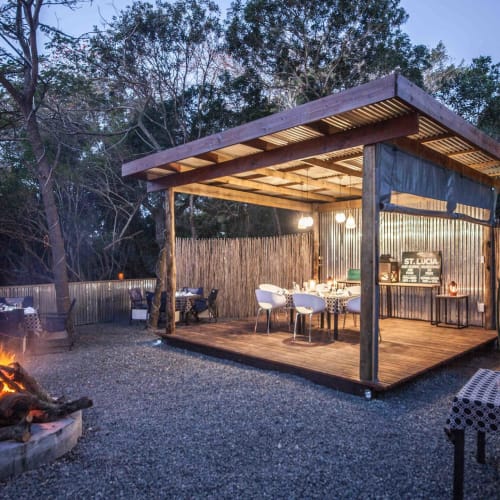 An outdoor dining area with a wooden deck and a thatched roof, surrounded by a rustic fence, featuring a fire pit and tables set for dining.