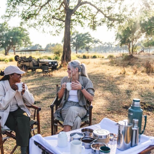 Two women enjoying coffee in an outdoor setting with trees and vehicles in the background.