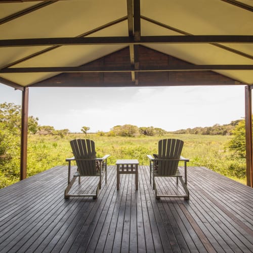 Two Adirondack chairs and a small table on a wooden deck overlooking a lush green landscape under a shelter.